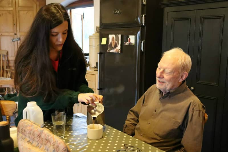 female live in carer pouring coffee for elderly man in cozy kitchen new age care